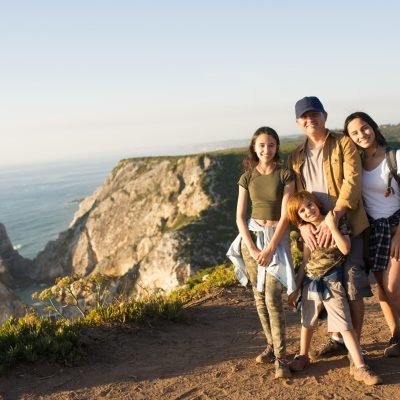 Happy father hiking with children in mountains. Portrait of mid adult man posing with teen daughters and preteen son against beautiful seascape. Active family weekend concept